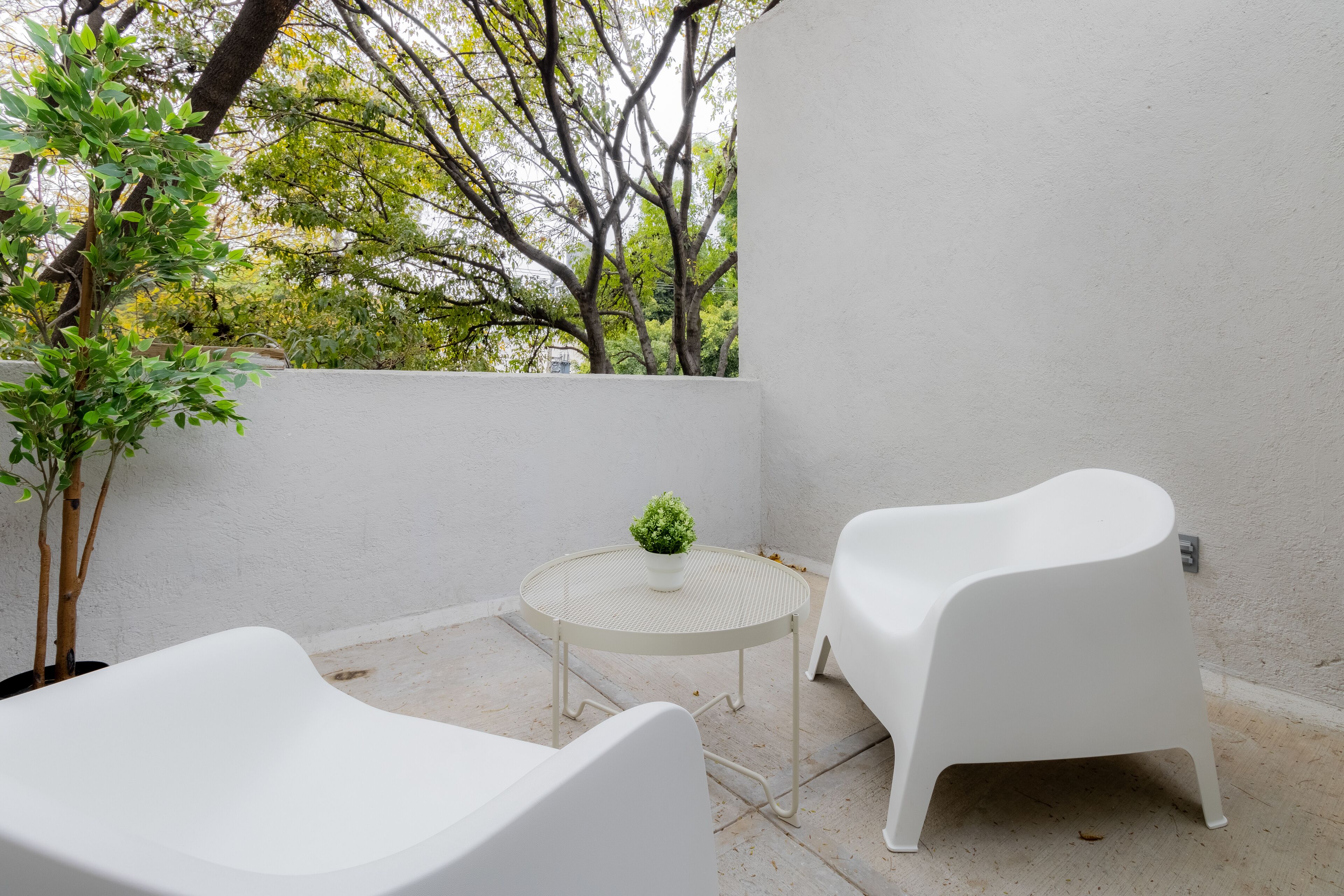 Un pequeño espacio al aire libre con dos sillas blancas y una mesa redonda, rodeado por un muro blanco y un árbol.