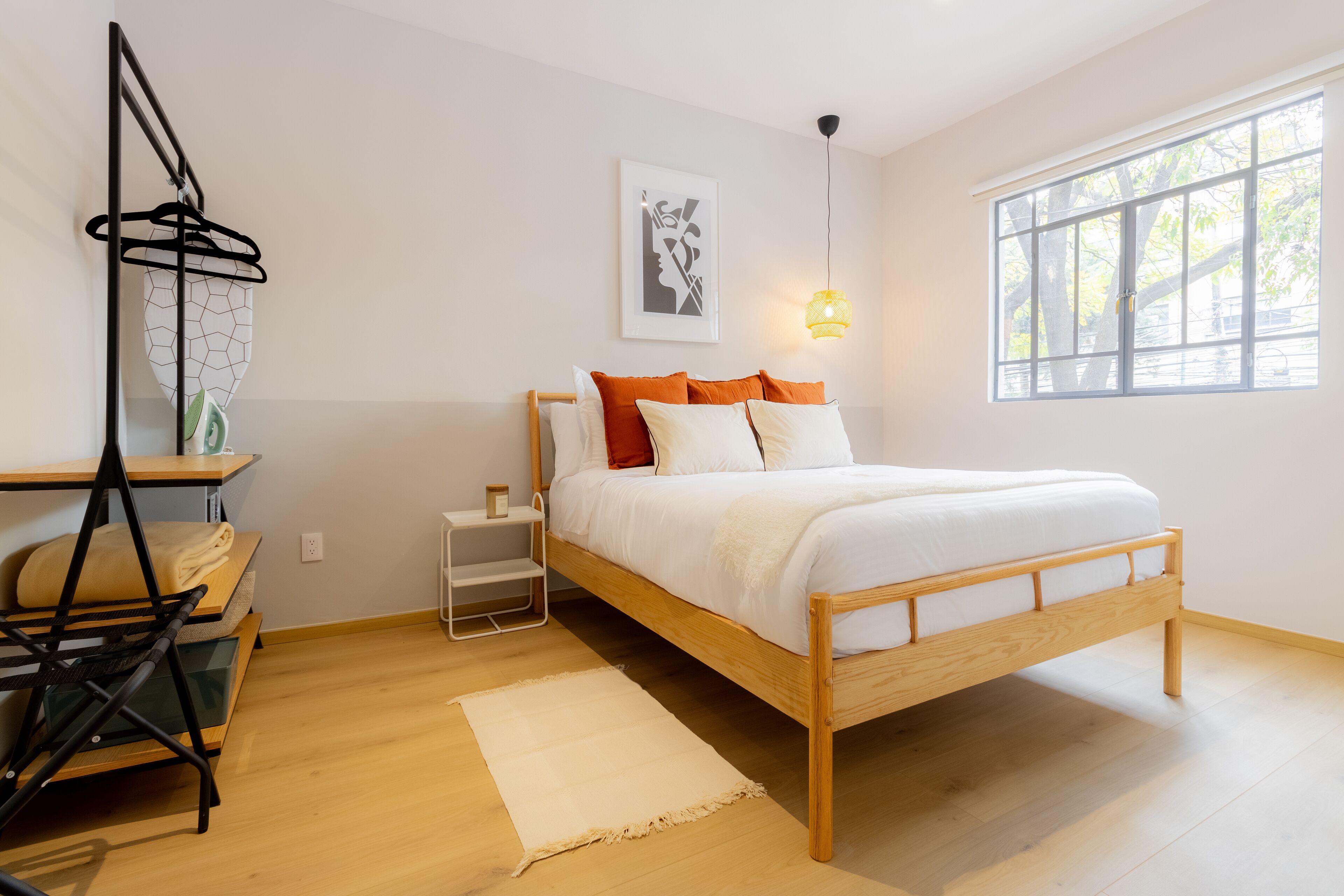 A bedroom with a wooden bed, white bedding, a small bedside table, a chair, a hanging light fixture, and a window with a view of trees.