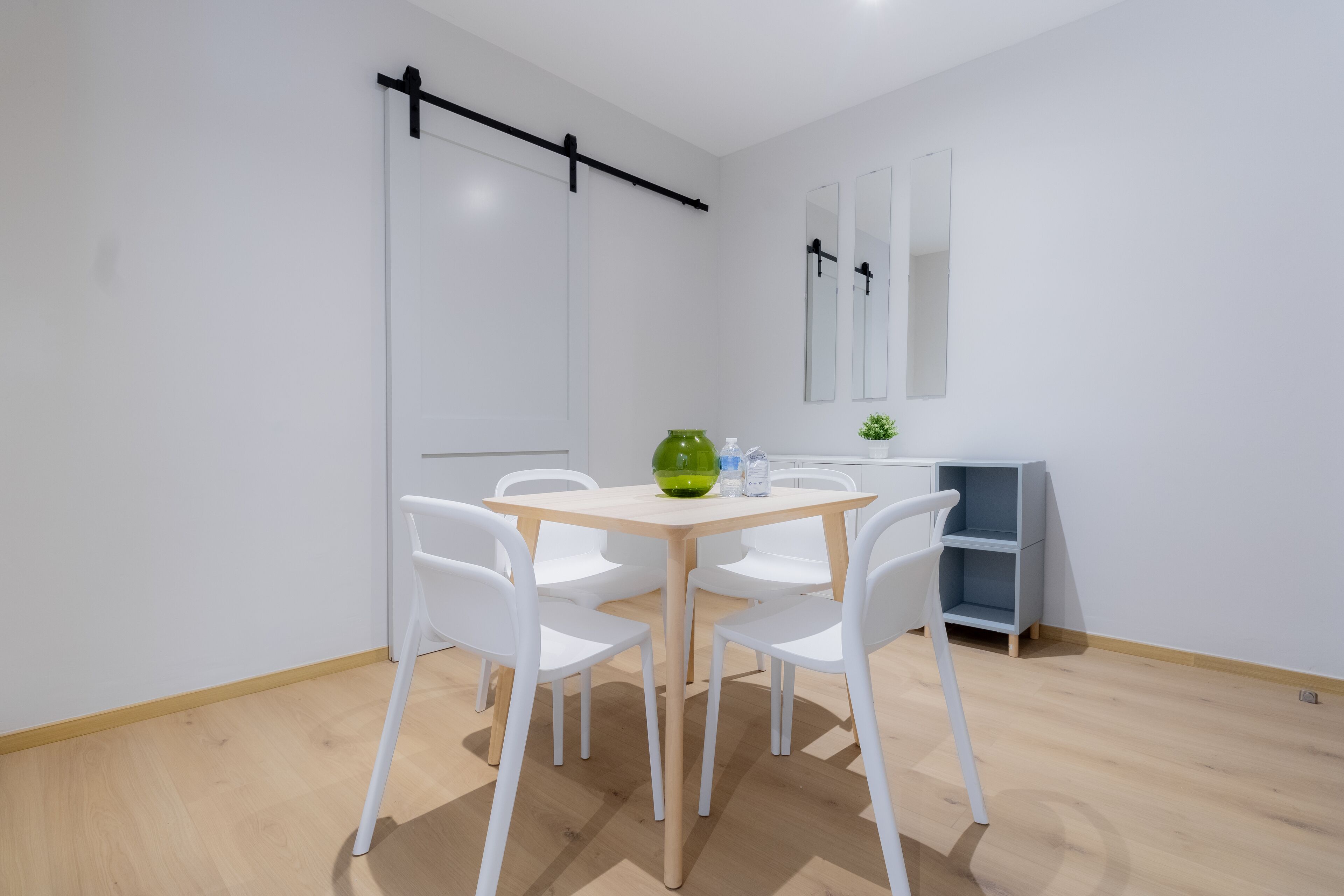 A modern, minimalist room with a wooden dining table and white chairs, a small green vase, and a mirror on the wall.