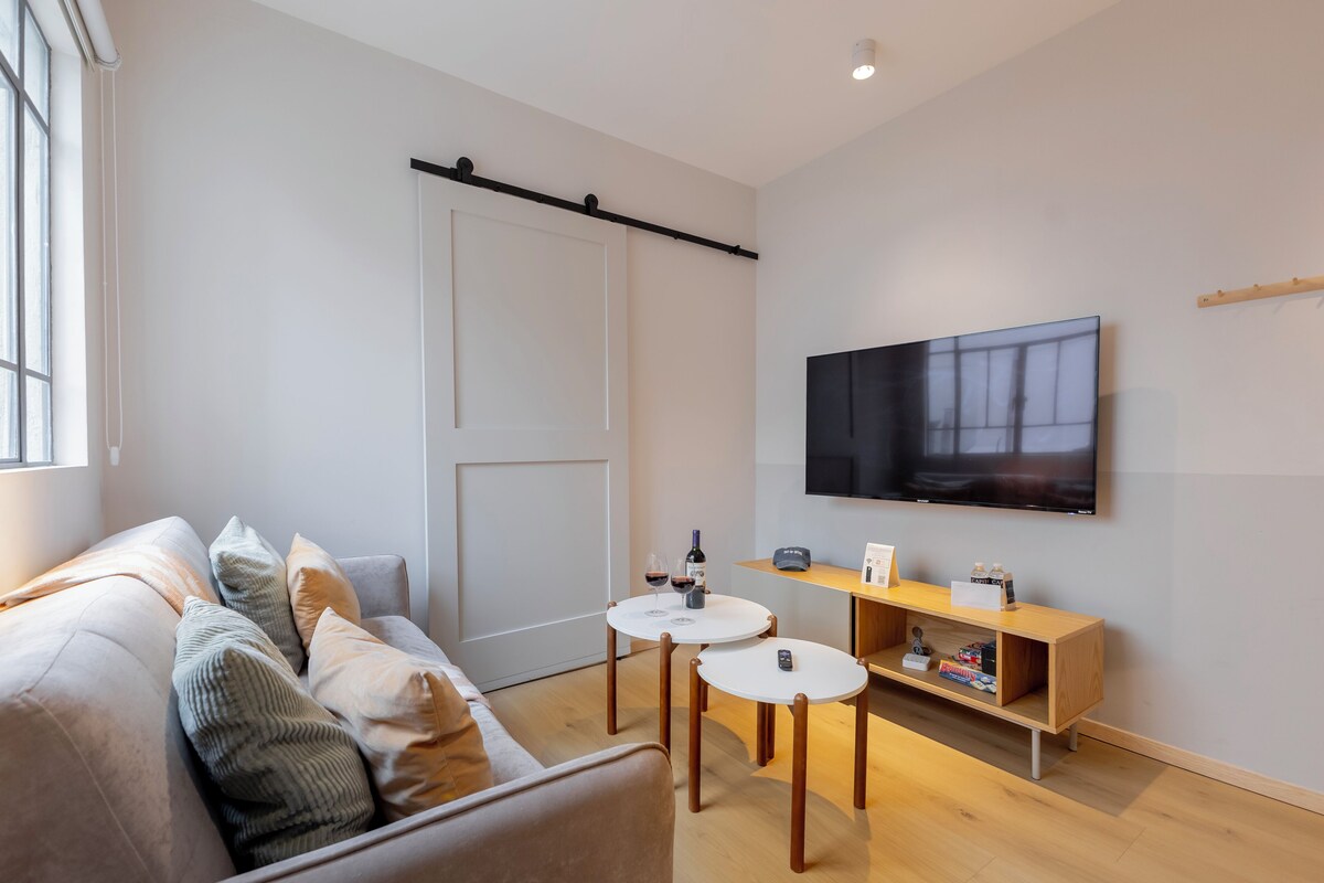 Una sala de estar moderna con un sofá, dos mesas redondas pequeñas, un televisor montado en la pared y un estante de madera con varios objetos.