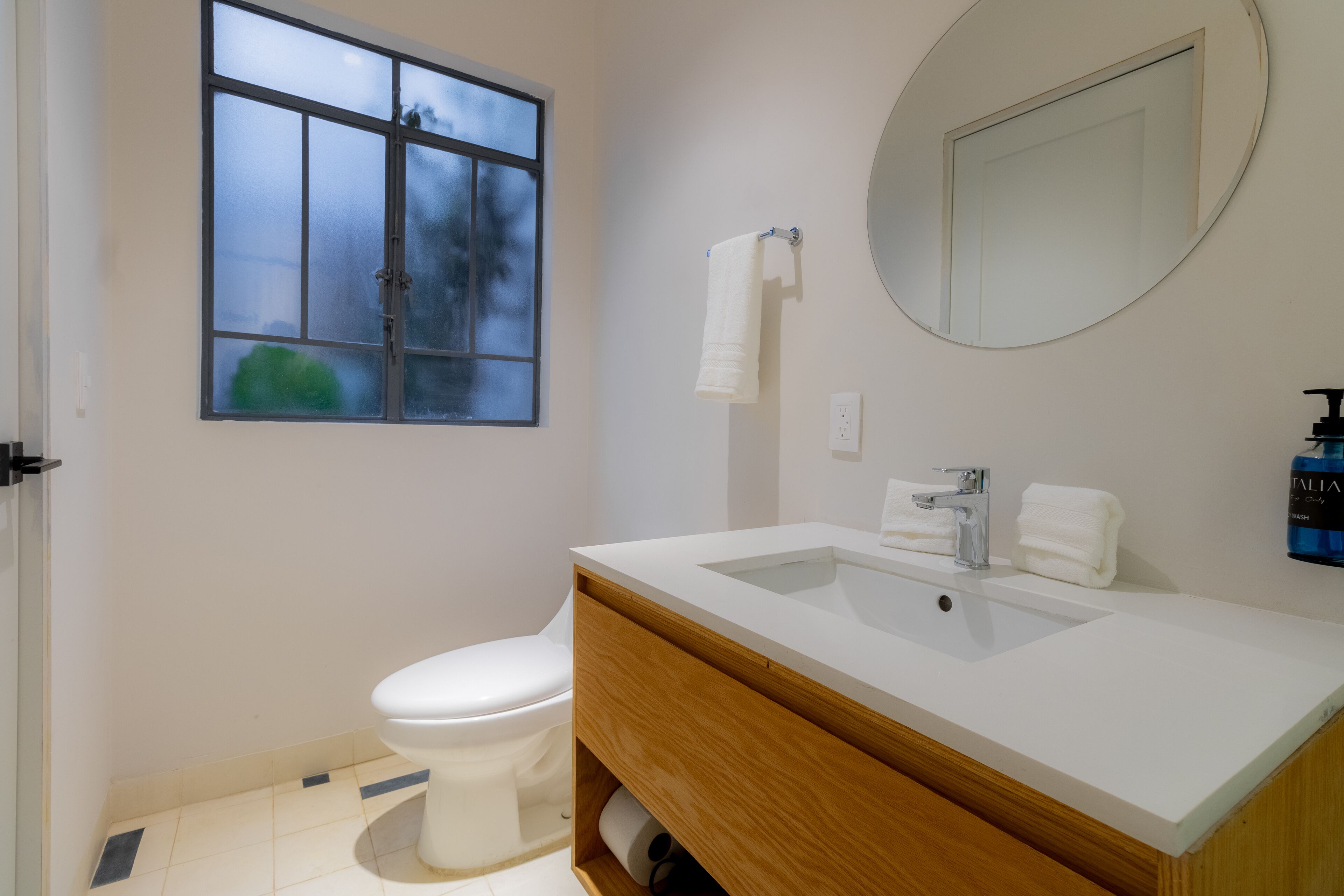 Ein Badezimmer mit weißem Waschbecken, einem Spiegel, einer Toilette und einem Fenster.