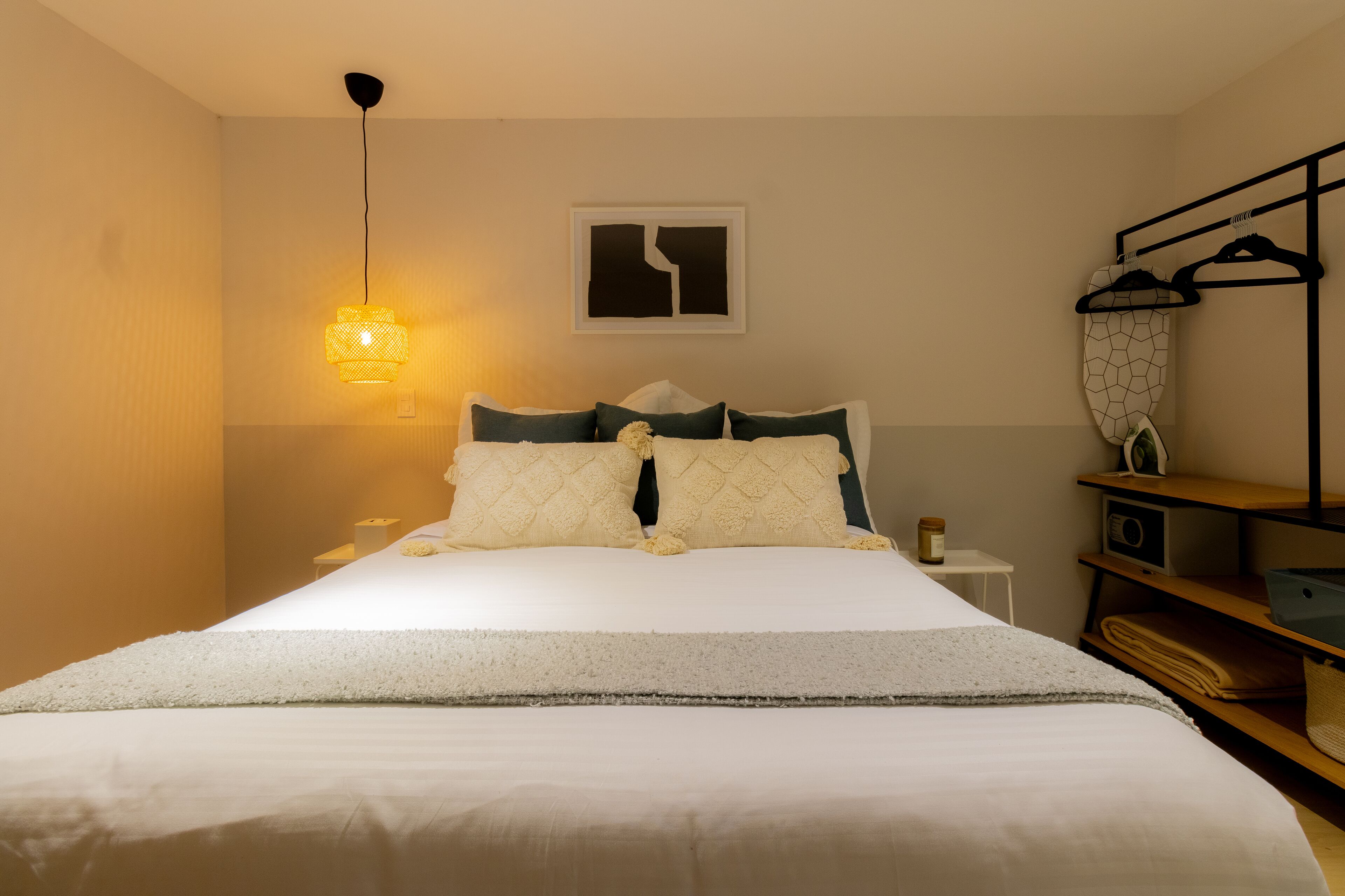 A neatly made bed with a white comforter and decorative pillows, a bedside lamp, a small table with a lamp, and a wall-mounted shelf with a radio and folded clothes.