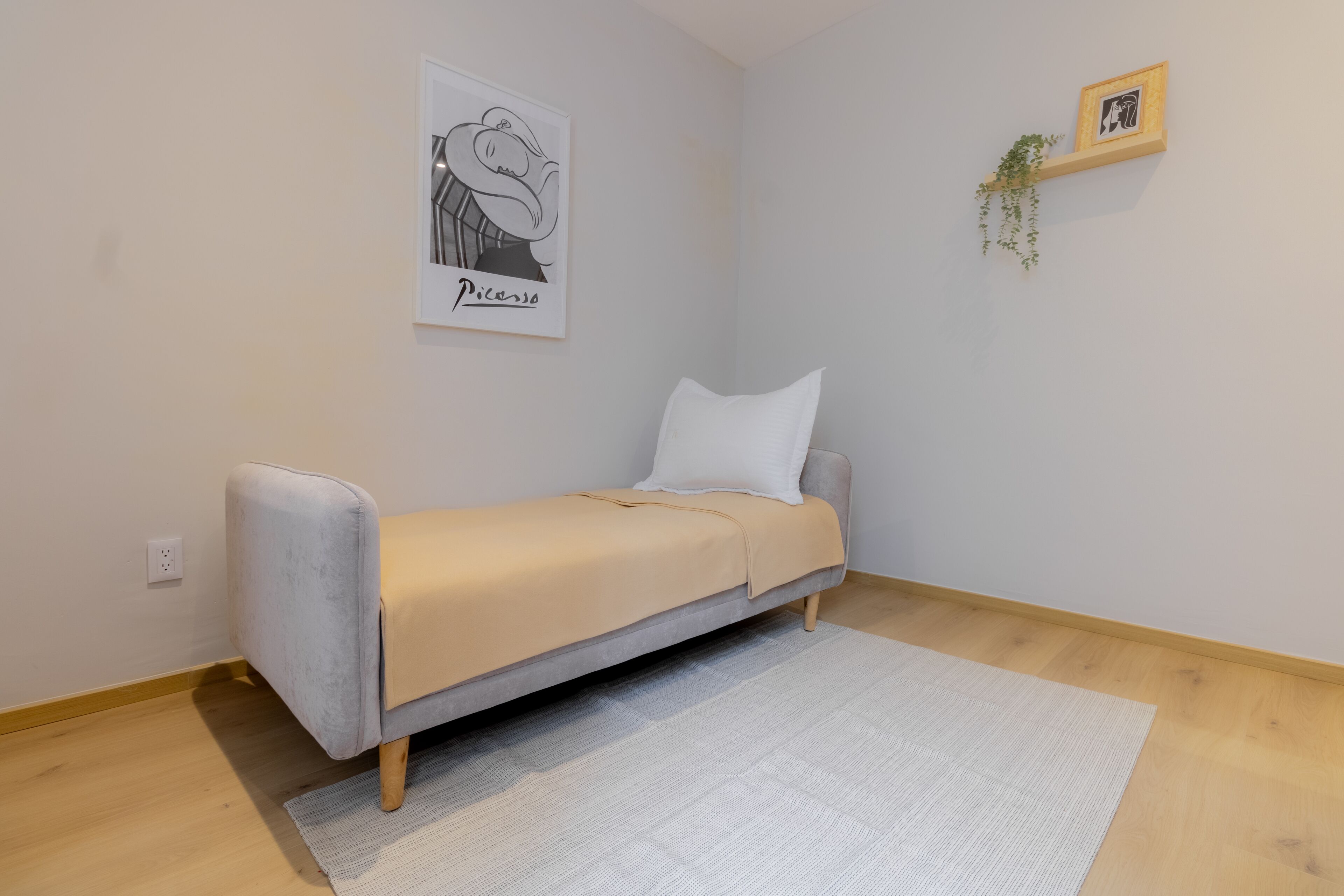 Ein kleiner, moderner Raum mit einem Einzelbett, einem hellen Teppich, einem gerahmten Kunstwerk an der Wand und einer kleinen Pflanze auf einem Regal.