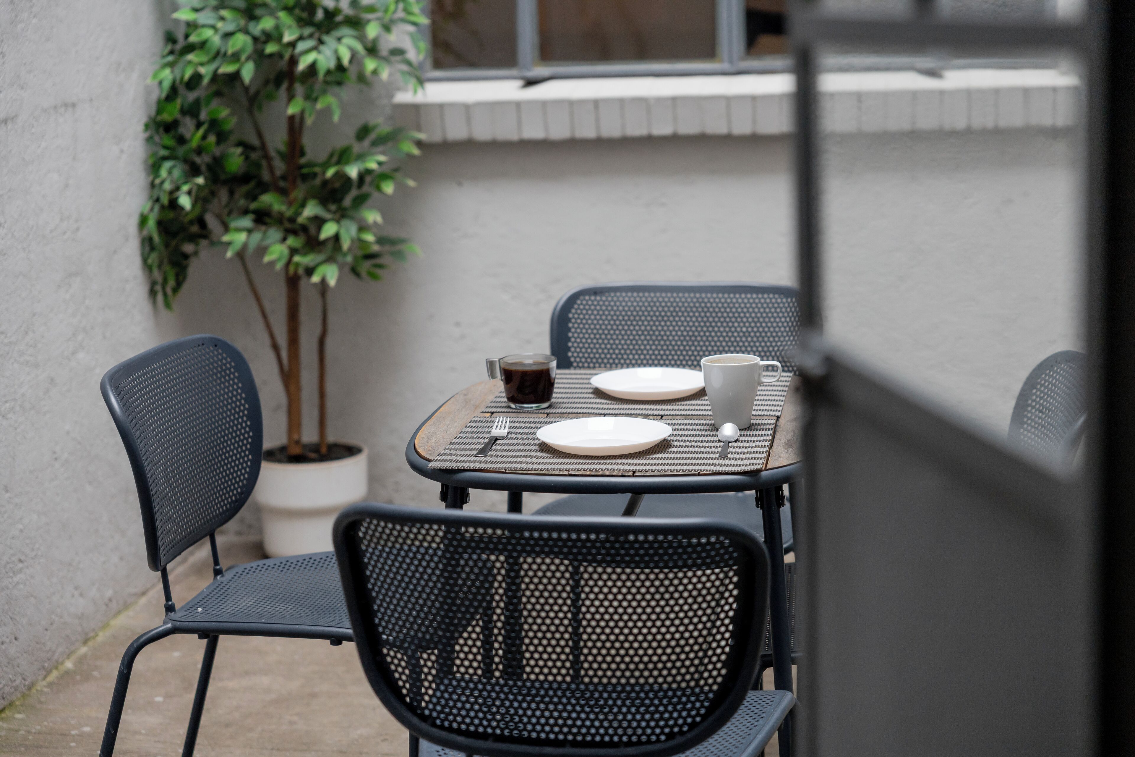 A small outdoor dining area with a table and chairs, a potted plant, and a window in the background.