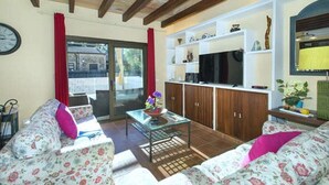 Ein Wohnzimmer mit floralen Sofas, einem Glastisch, Holzschränken und einer Schiebetür zu einem Außenbereich.