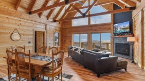 A log cabin interior with a dining area, sofa, fireplace, and large windows. - Custom-Built Log Cabin with Long Range Views (Banner Elk)