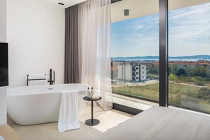 A modern bathroom with a freestanding bathtub, a round table with toiletries, and a large window offering a city view. - 22 (Zadar)