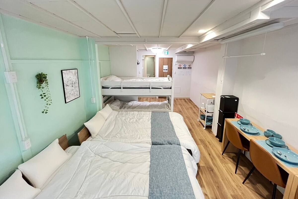 A dormitory room with bunk beds, a desk with chairs, and a small kitchenette area.