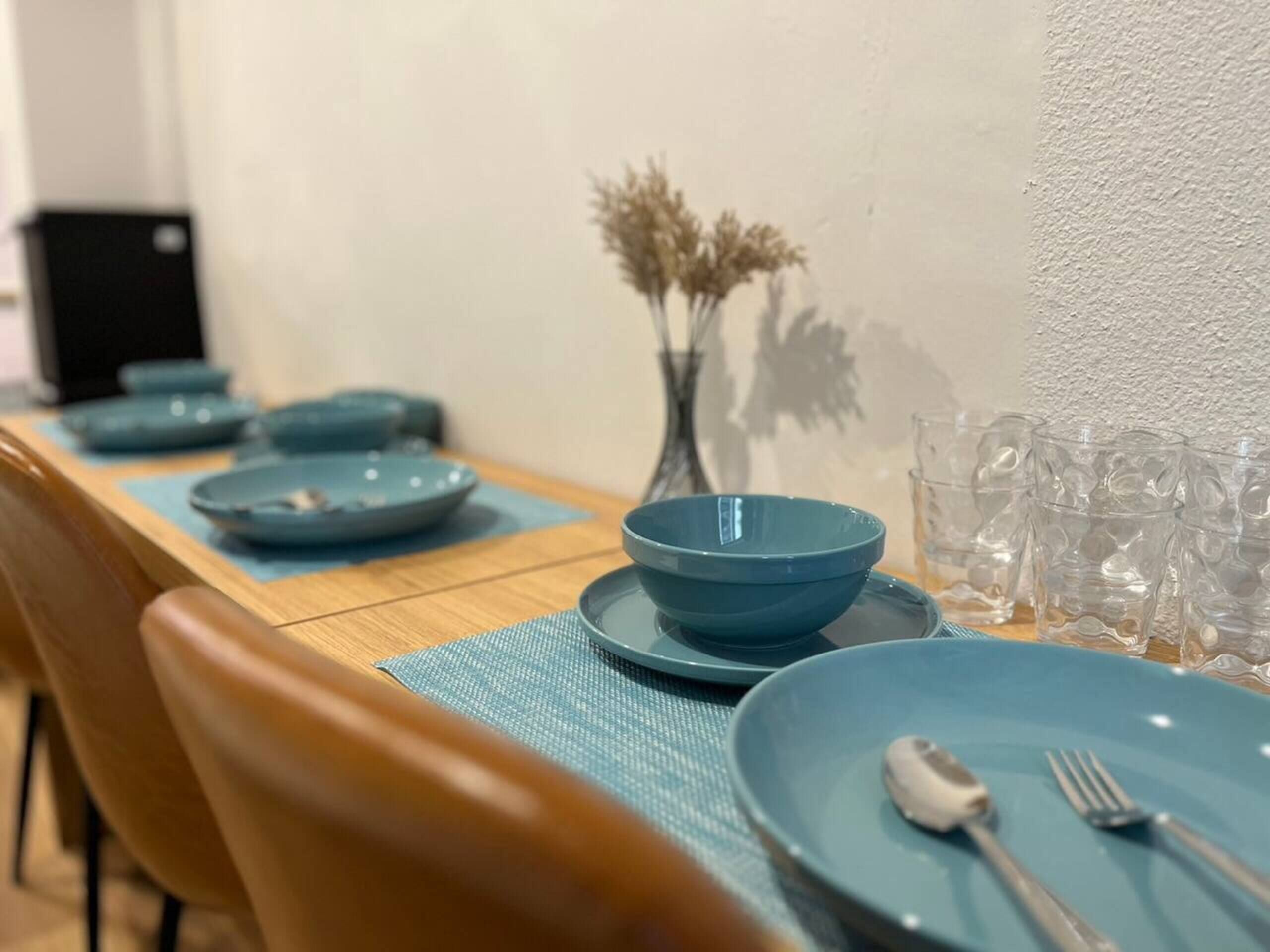 Une table à manger dressée avec de l’assiette, des bols et des couverts bleus, agrémentée d’un vase rempli de fleurs séchées.