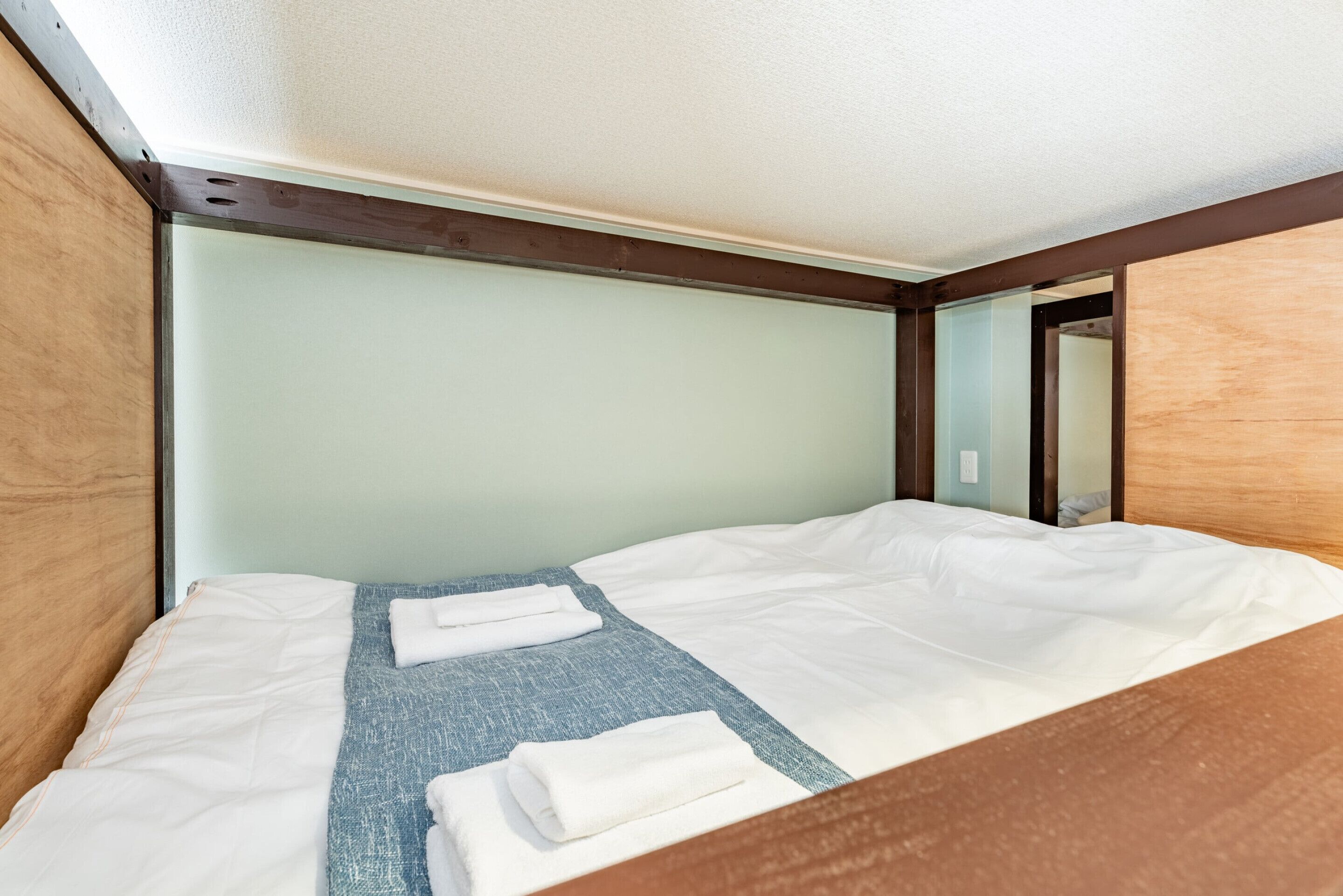 A single bed with white bedding, a blue blanket, and two folded white towels. The room has wooden walls and a wooden headboard.