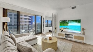 A modern living room with a large flat-screen TV, a beige sofa, and a round coffee table. - Cute Ocean View Condo Beach Service 1217 (Miami Beach)