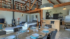 Un salón espacioso con techo alto, vigas de madera a la vista, una chimenea, una mesa de comedor para cuatro comensales y una cocina al fondo.