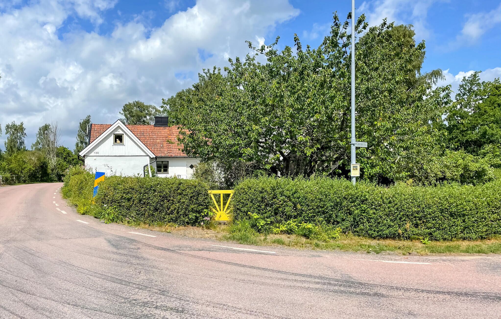 A rural road with a white house, dense greenery, and a yellow traffic sign.