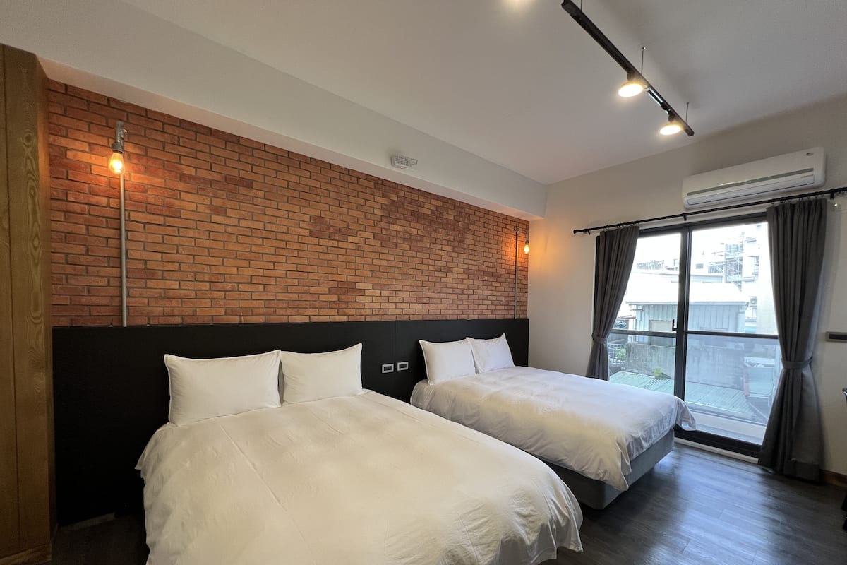 A hotel room with two beds, a brick wall, and a large window with curtains.