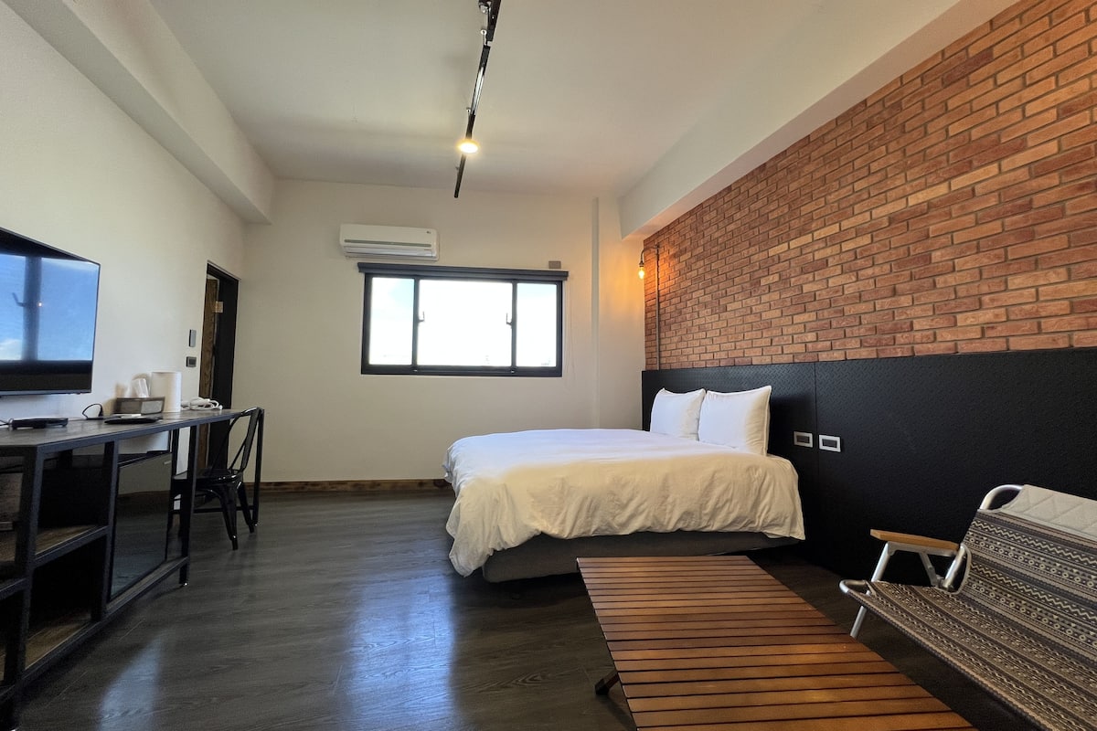A hotel room with a bed, a desk with a chair, a television, a window, and a brick wall.