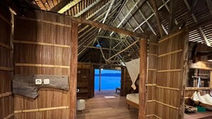 A wooden room with a bed, a bench, and a table. There is a view of the sea through an open doorway. - Wai Resort - Raja Ampat  (Jerief Island)