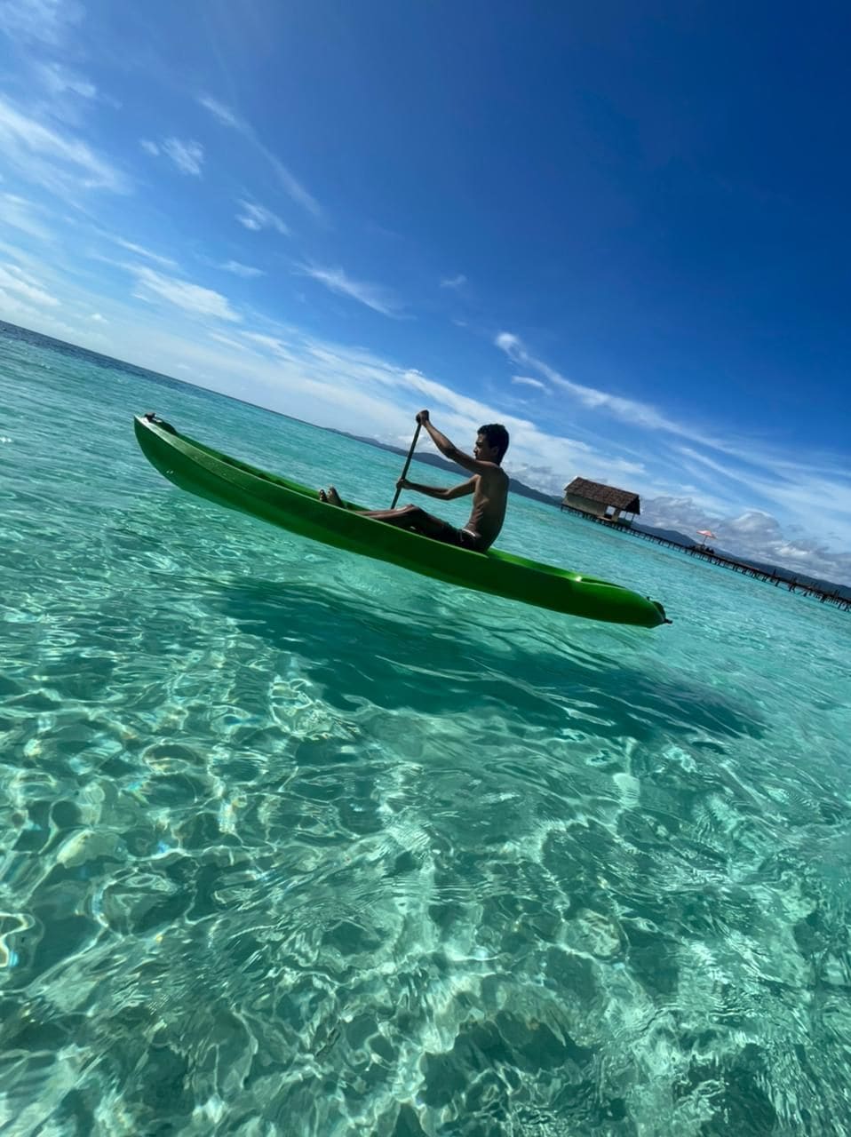 A person paddling a green canoe on clear turquoise water with a wooden structure in the background.