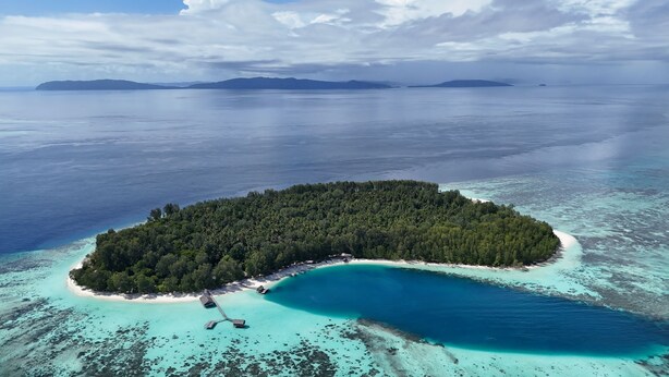 An aerial view of a tropical island with a clear turquoise lagoon, surrounded by a dense forest and a small beach area with overwater bungalows. - Wai Resort - Raja Ampat  (Jerief Island)