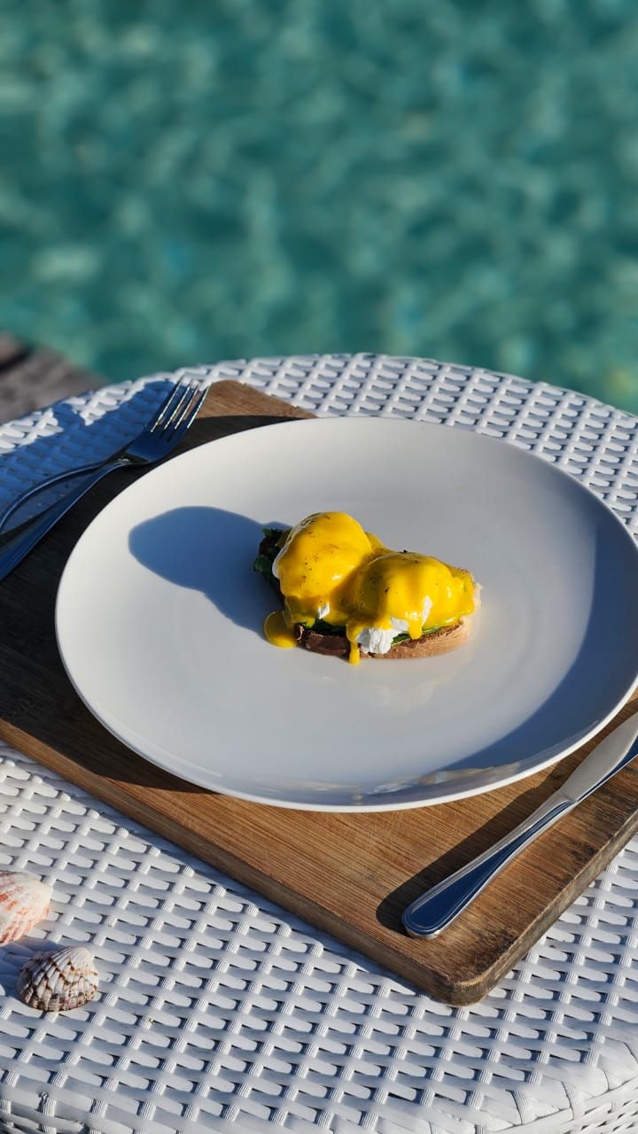 A plate of eggs benedict on a white plate with a wooden board underneath, placed on a white table with a blue background.