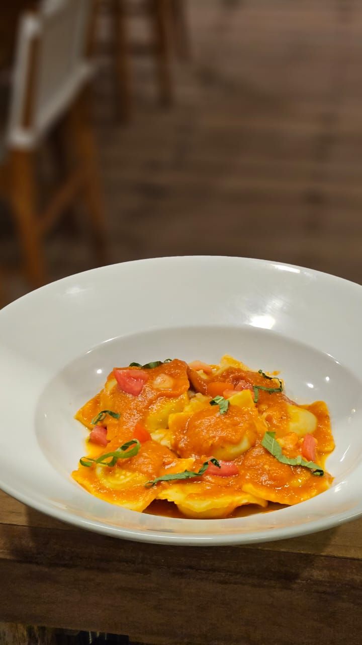 A white plate with a dish of ravioli in a tomato-based sauce, garnished with herbs, on a wooden table.