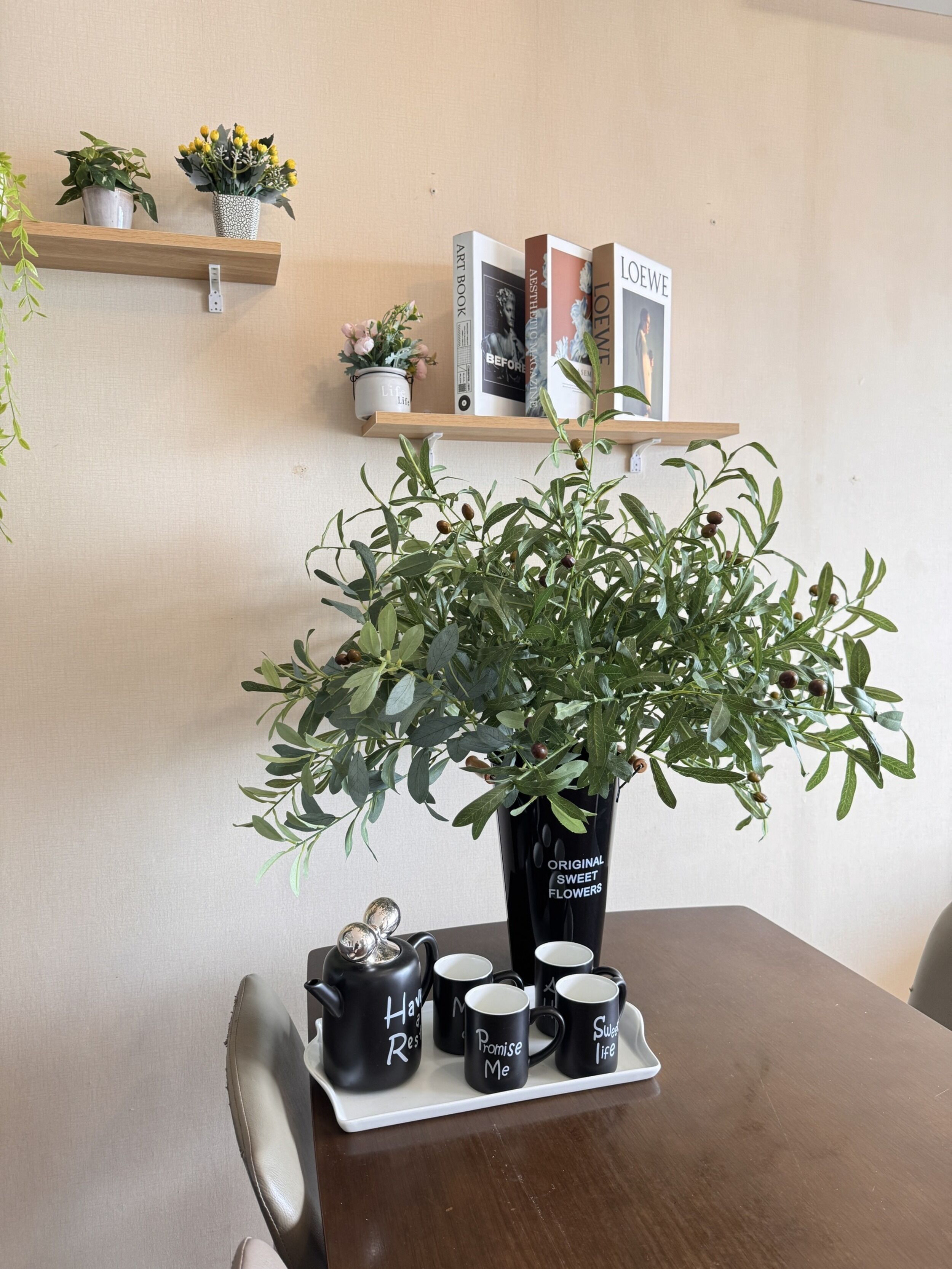 A table with a black vase of 'Original Sweet Flowers', a tea set, and 'Promise Me' mugs. There are books on a shelf above and a potted plant to the left.