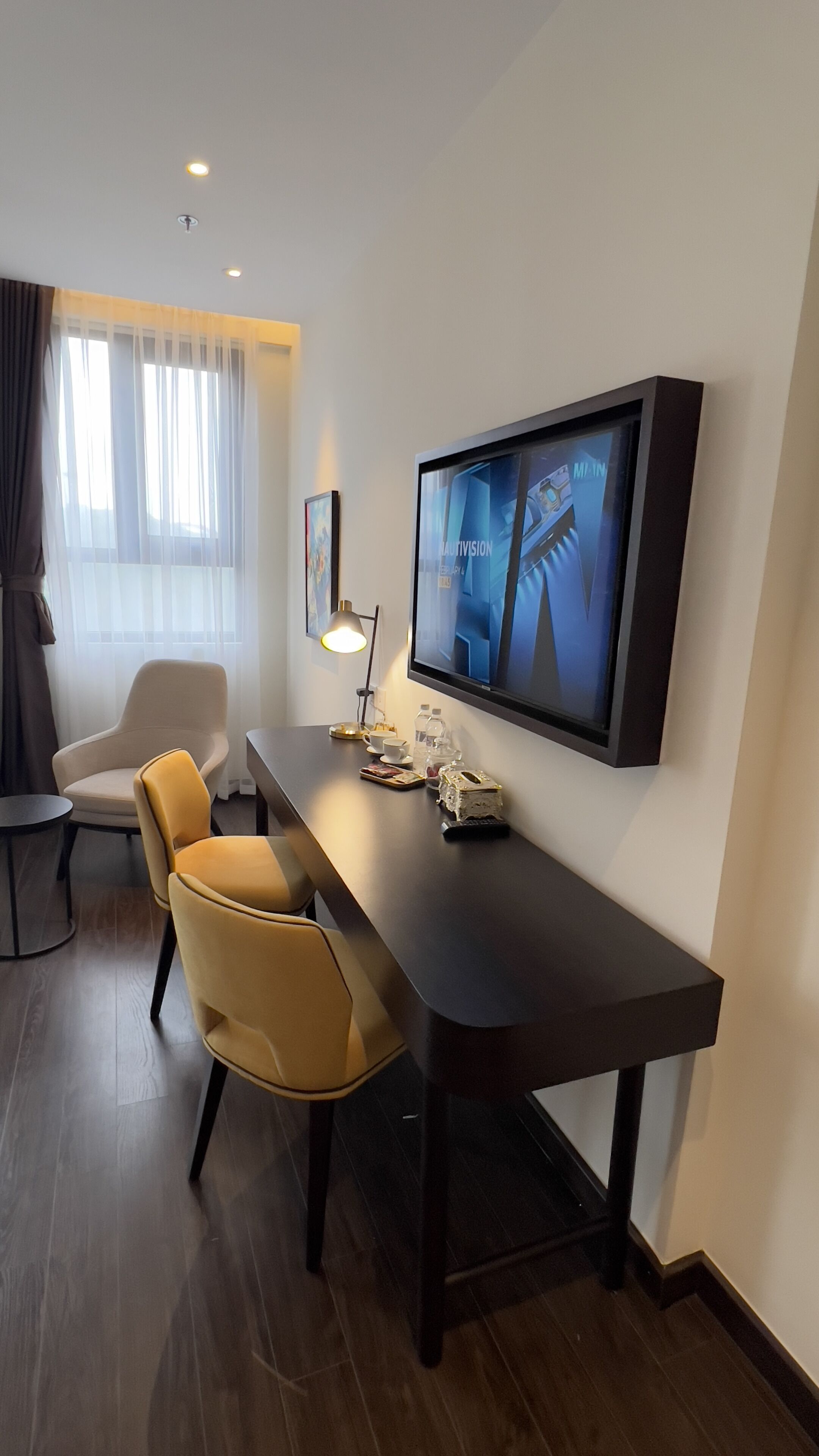 A modern hotel room with a dark wooden desk, a comfortable chair, a flat-screen TV mounted on the wall, and a window with sheer curtains.