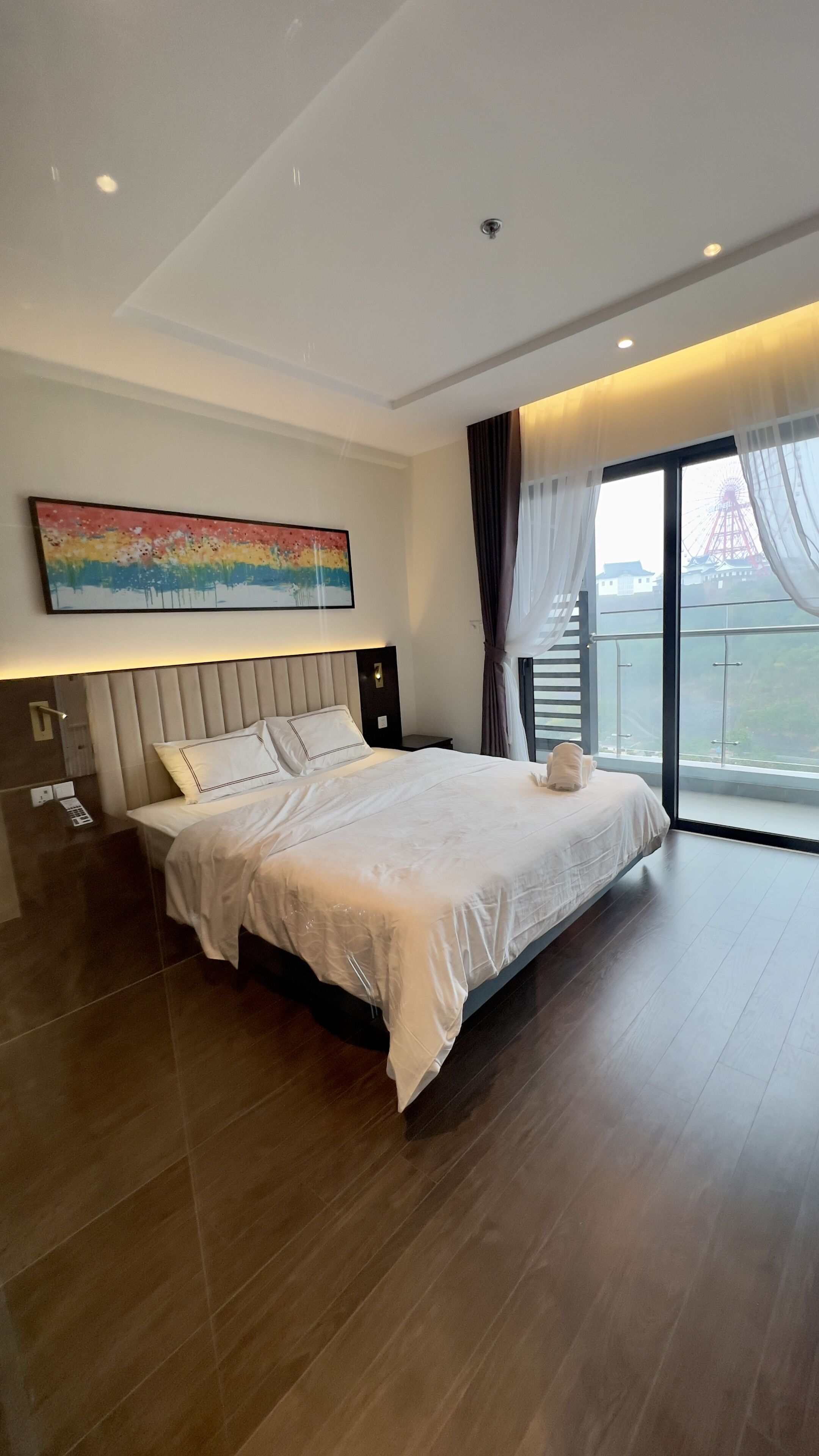 Een moderne hotelkamer met een groot bed, een nachtkastje, een schilderij en uitzicht op een reuzenrad door het raam.