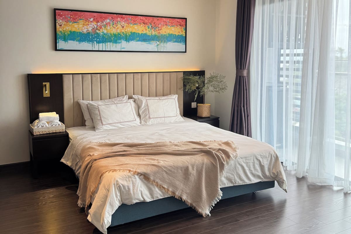 A modern hotel room with a large bed, a headboard, a bedside table with a lamp, a wall-mounted painting, and a large window with curtains.