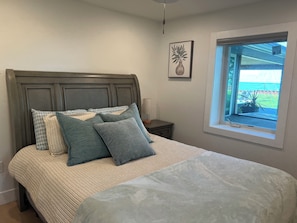 A bedroom with a bed, pillows, a nightstand, a lamp, a framed picture, and a window with a view. - Newly Renovated Waterfront Cottage Lake St. Clair (Pointe aux Roches)