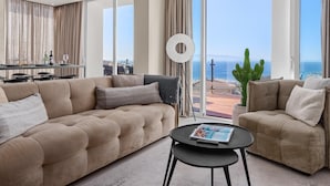 A spacious living room with a large sofa, a smaller sofa, and a coffee table. There is a dining area with a table and chairs. The room has large windows with a view of the ocean and a cactus plant. - Karat Villa Sand Torviscas (Costa Adeje)