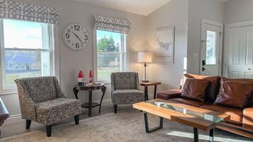 A living room with patterned armchairs, a glass-top coffee table, a brown leather sofa, a wall clock, and a framed map.
