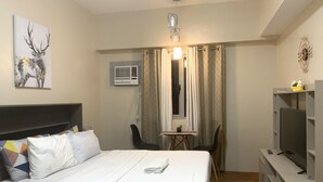 A hotel room with a bed, a desk, a chair, a TV, and an air conditioning unit.