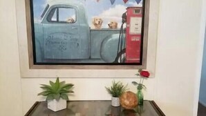 A framed painting of a blue truck with two dogs and a gas pump on a wall. Below the painting, there is a glass-top table with two potted plants and a vase with a red rose. - River House (Parker)