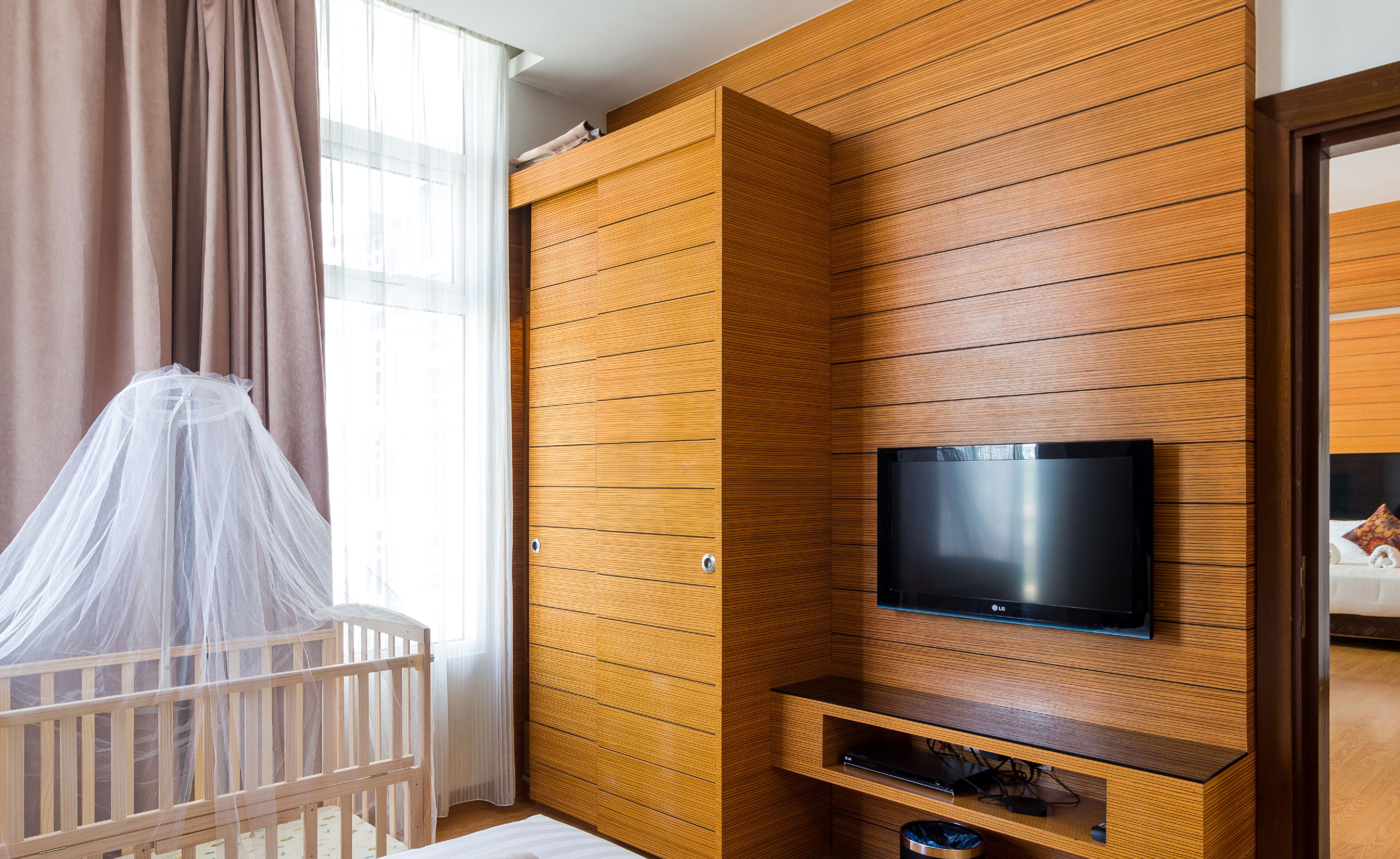 Une pièce avec un mur lambrissé en bois, un téléviseur fixé au mur, un berceau surmonté d’un dais blanc et une fenêtre ornée de rideaux légers.
