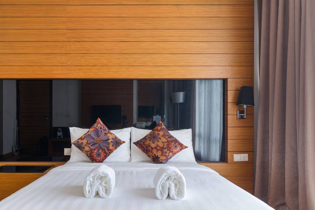 Une chambre d’hôtel avec un grand lit, une tête de lit en bois, deux coussins décoratifs, une table de chevet, un miroir et une fenêtre avec des rideaux.
