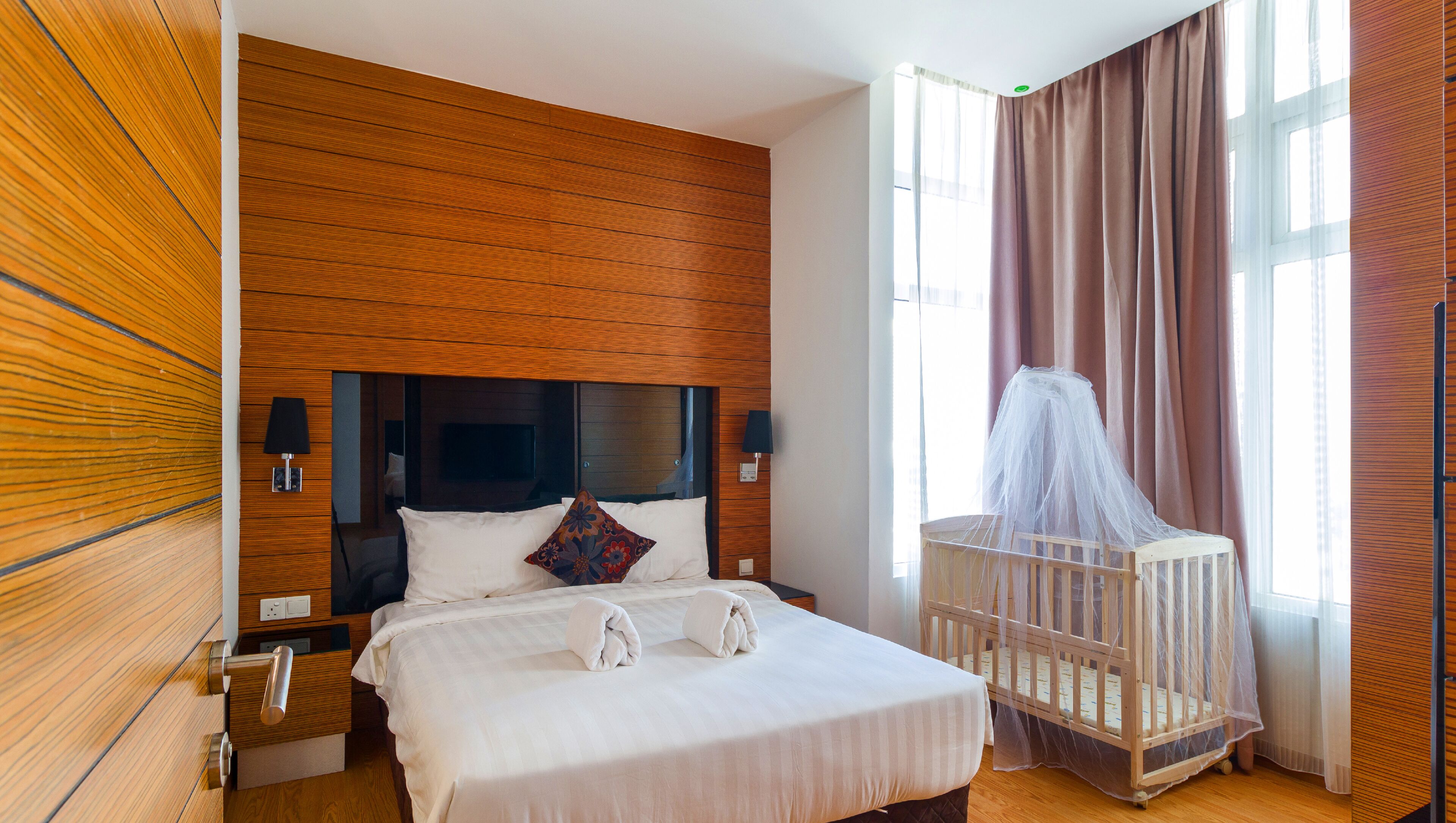 A hotel room with a wooden headboard, a bed with white linens, a bedside table with a lamp, a crib with a canopy, and a large window with curtains.