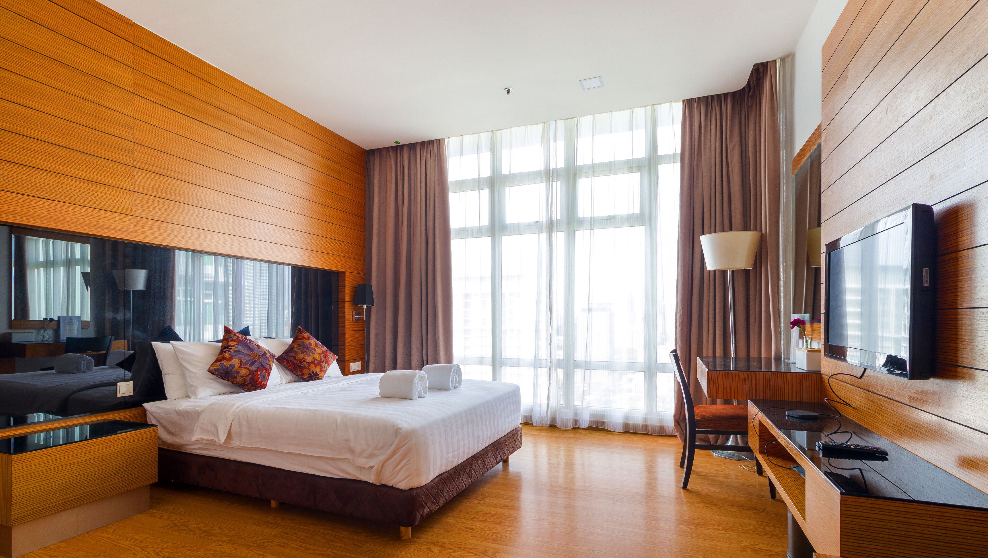 A modern hotel room with a large bed, wooden panel walls, a desk with a lamp, and a TV mounted on the wall.