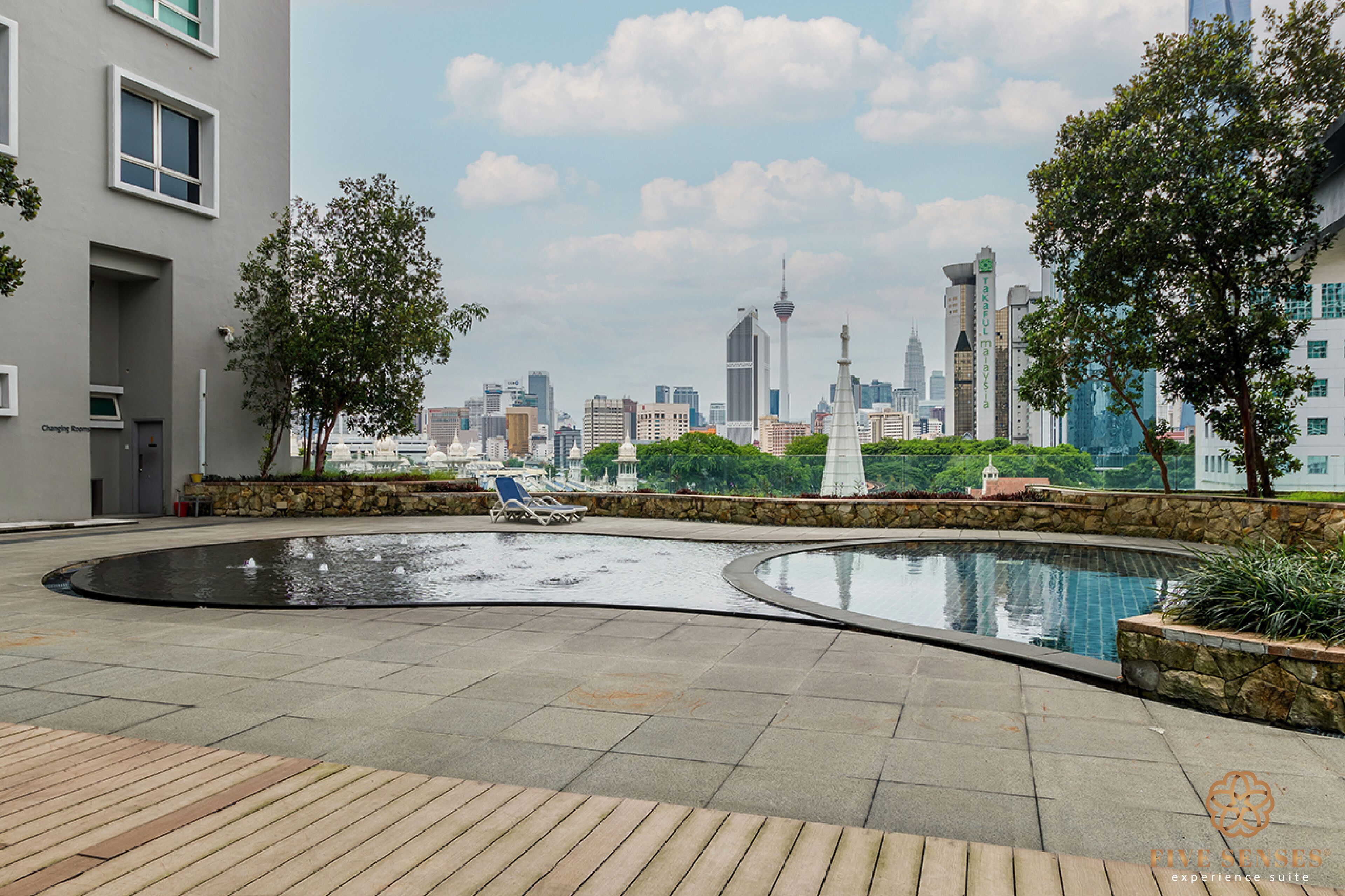 A rooftop pool area with lounge chairs, a stone wall, and a city skyline in the background.