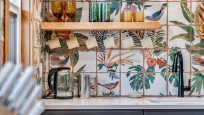 Een keuken met een betegelde spatwand met tropische bladmotieven, een houten plank met serviesgoed en een koffiezetapparaat.
