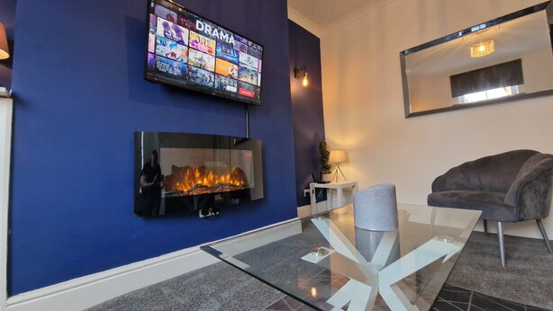 A modern living room with a blue accent wall, a flat-screen TV displaying a movie channel, a fireplace, a glass coffee table, and a comfortable sofa. - 2-Bed Home - Sleeps 4, Parking & Garden (Darlington)