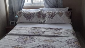 A neatly made bed with patterned bedding, a bedside table, and a window with curtains. - Kasoa Apartment with 2 Bedrooms (KASOA)