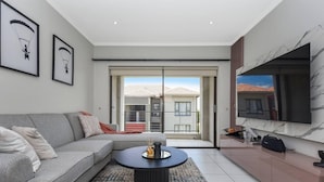 A modern living room with a grey sofa, a coffee table, and a TV on a marble wall. - 2 Bed Flat - Sleeps 4 - Pool - Garden - Parking (Sandton)