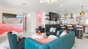 A modern living room with teal sofas, a pink accent wall, and a kitchen area with black cabinets and gold pendant lights.