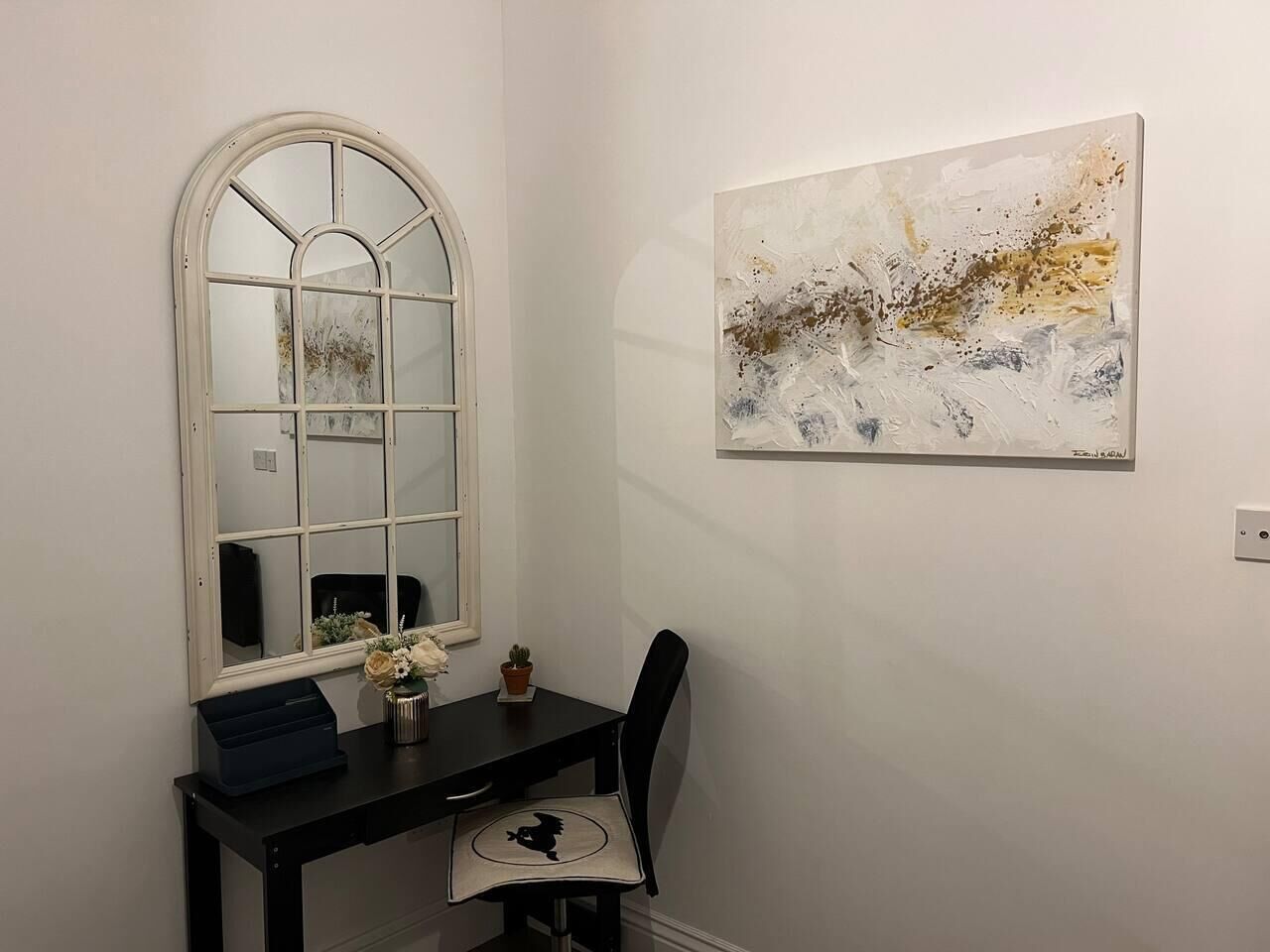 A room with a desk, chair, and a large window with a mirror. There is a painting on the wall and a small potted plant on the desk.