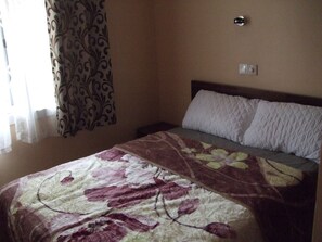 A bedroom with a bed, a nightstand, a wall-mounted light, and a window with curtains.