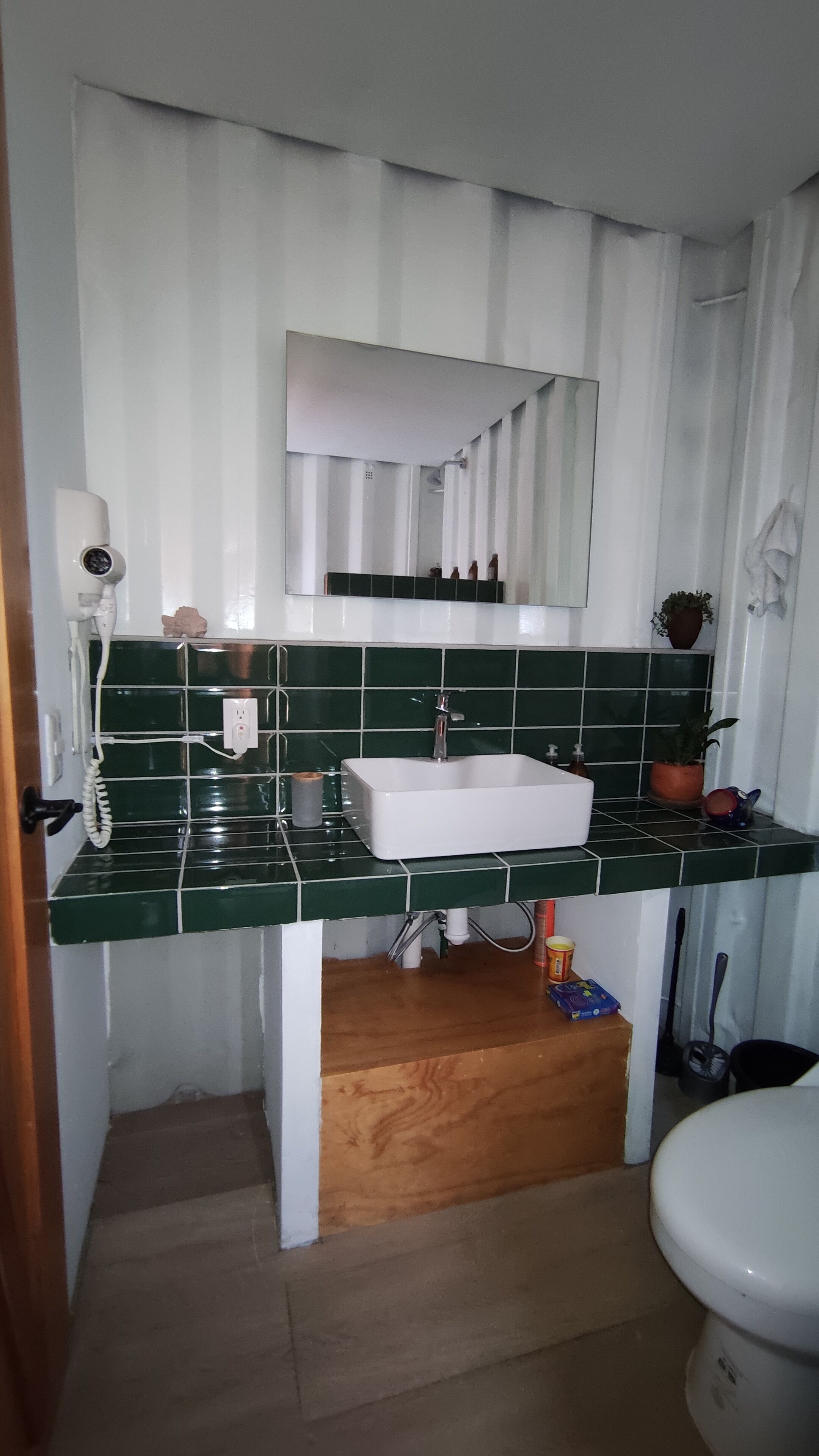 Un bagno con lavandino bianco, schienale rivestito in piastrelle verdi, uno specchio sopra il lavandino, un water e un mobiletto in legno sotto il lavandino.