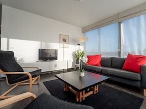 Una sala de estar moderna con un sofá negro, una mesa de centro de madera, un sillón negro y un televisor sobre un mueble blanco.