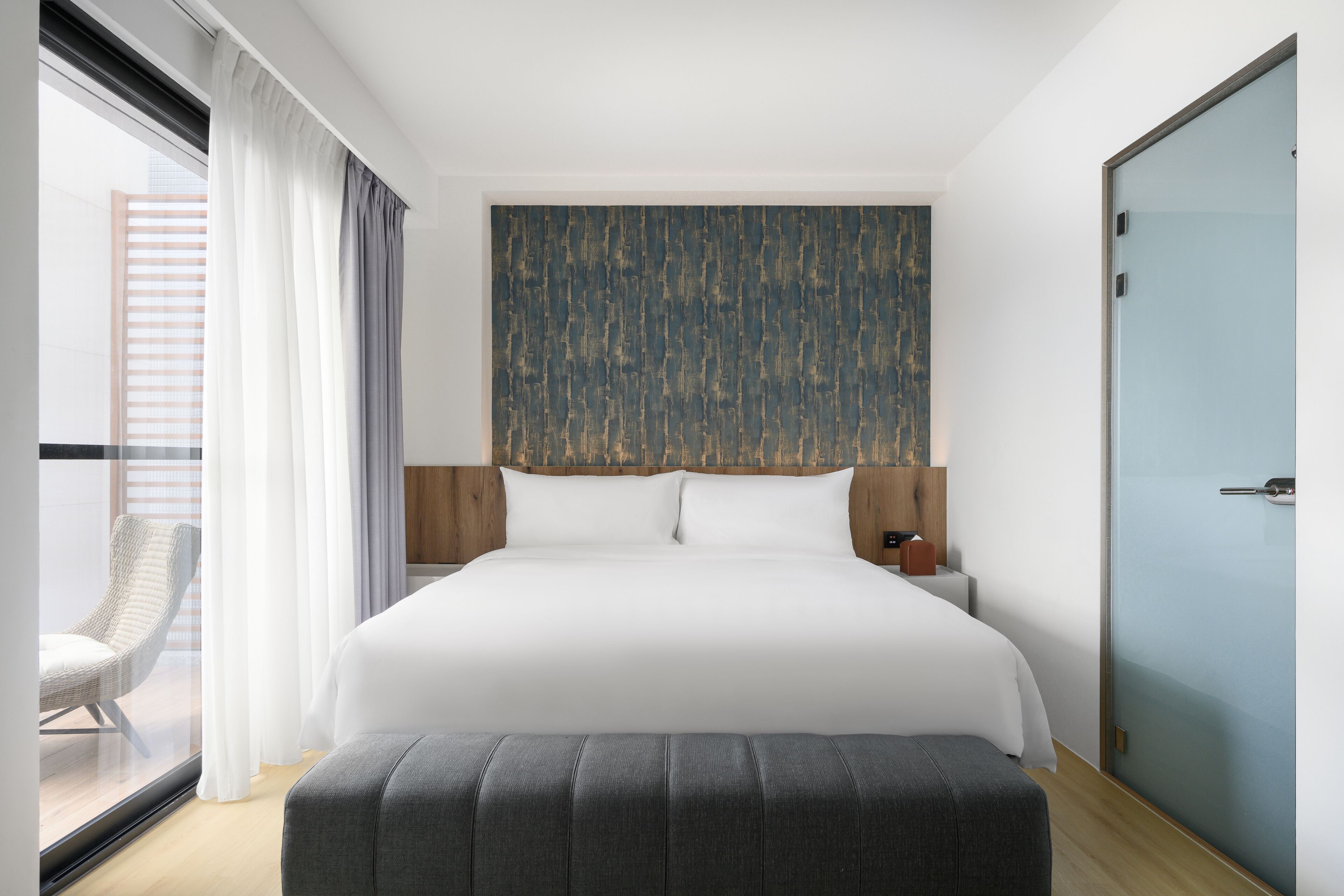 A modern hotel room with a large bed, a grey upholstered bench, a wooden headboard, a glass door, and a view of the city through the window.