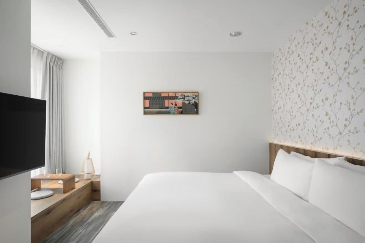A modern hotel room with a large bed, a flat-screen TV, a wooden side table, and a wall-mounted artwork.