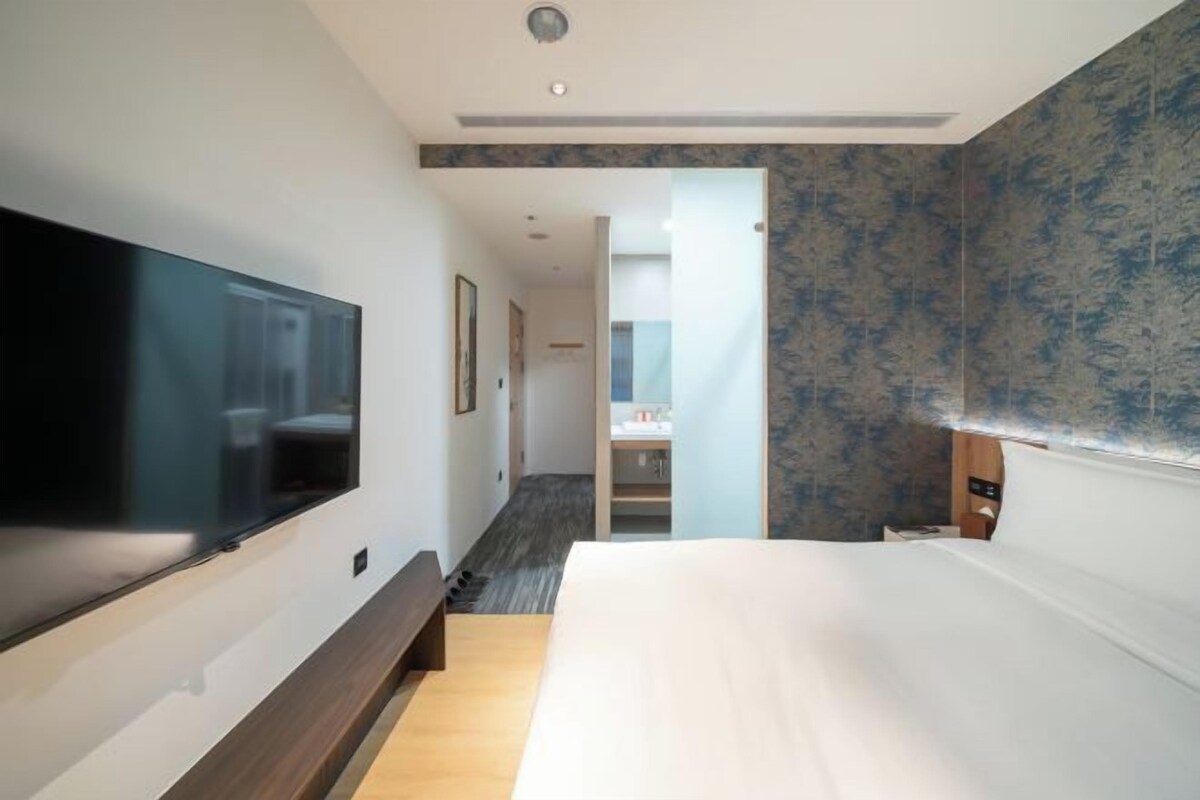 A modern hotel room with a large flat-screen TV mounted on the wall, a wooden bench, a large bed, and a bathroom visible through an open door.